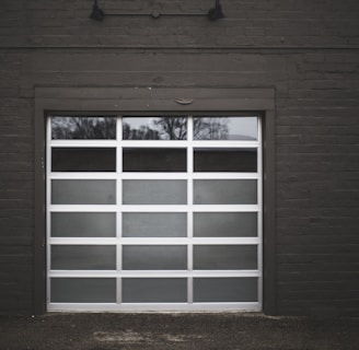 A modern white garage door with several rectangular glass panels is set within a dark brick exterior. The reflection of bare trees is visible in the glass panels, suggesting a winter setting.