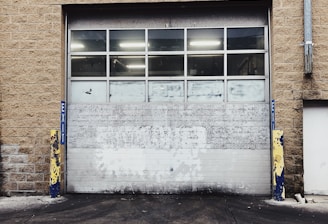 A large industrial garage door set into a tan brick building. The door is white with multiple small windows at the top, and there are yellow and blue protective bollards on either side. The ground in front of the door is paved and appears slightly worn.