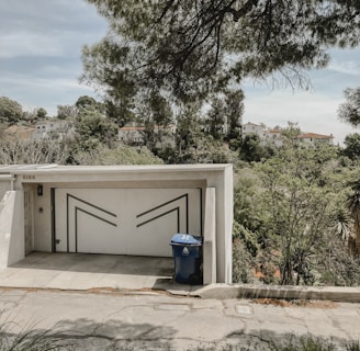 A modern garage door with geometric patterns stands at the end of a short driveway. A blue recycling bin is positioned in front of the garage. Surrounding the building, lush greenery and tall trees extend into the background, with hillside houses visible beyond the foliage.