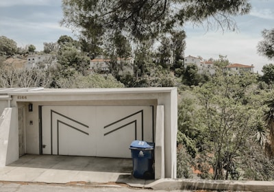 A modern garage door with geometric patterns stands at the end of a short driveway. A blue recycling bin is positioned in front of the garage. Surrounding the building, lush greenery and tall trees extend into the background, with hillside houses visible beyond the foliage.